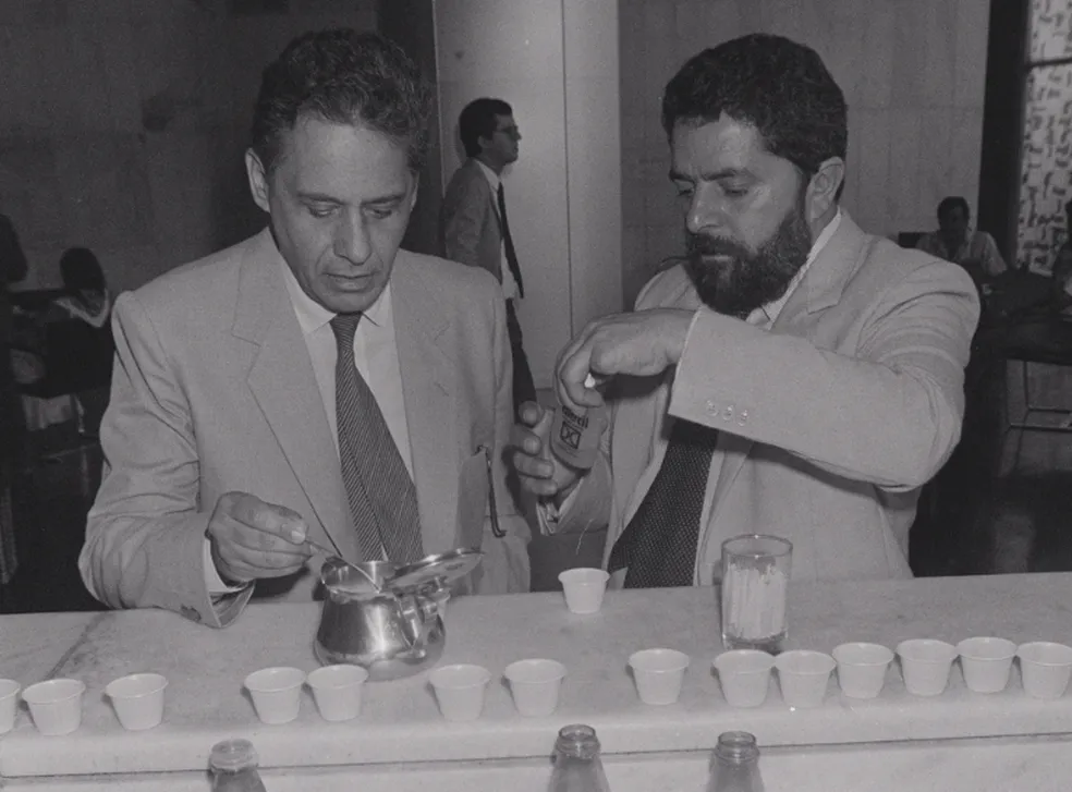 Lula e FHC em frente a um balcão cheio de copinhos descartáveis, adoçando o que parece ser seus cafés.